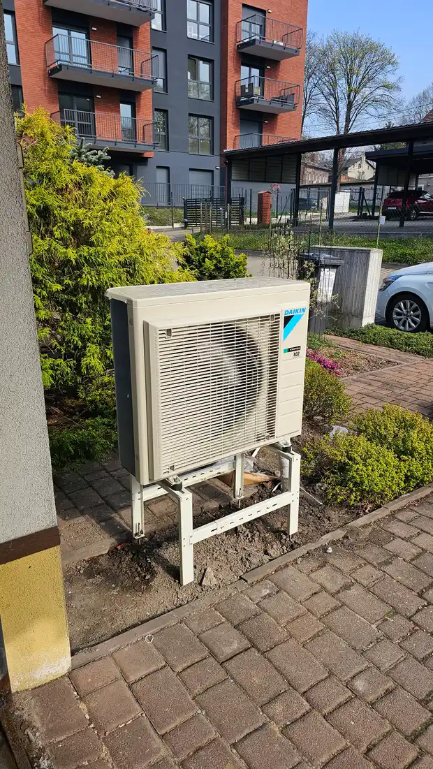 Jednostka zewnętrzna Daikin na białym stelażu przy chodniku i żywopłocie